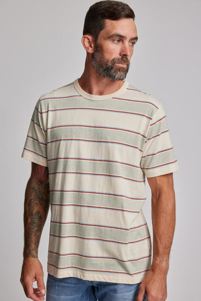 A man with short brown hair and a beard wears the Barkers Arnord Stripe Tee, a beige tee with red and gray block stripes, paired with blue jeans. He stands sideways against a plain background, revealing a tattoo on his left forearm.