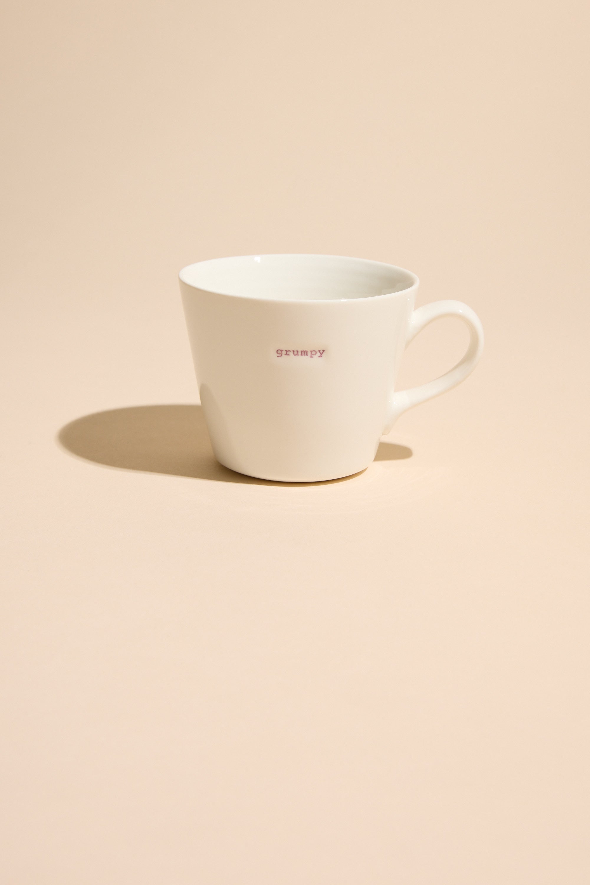 The Bucket Mug - Grumpy by Other is a white porcelain mug with “grumpy” in small pink letters on the front, shown on a light beige surface with a soft shadow to the left.