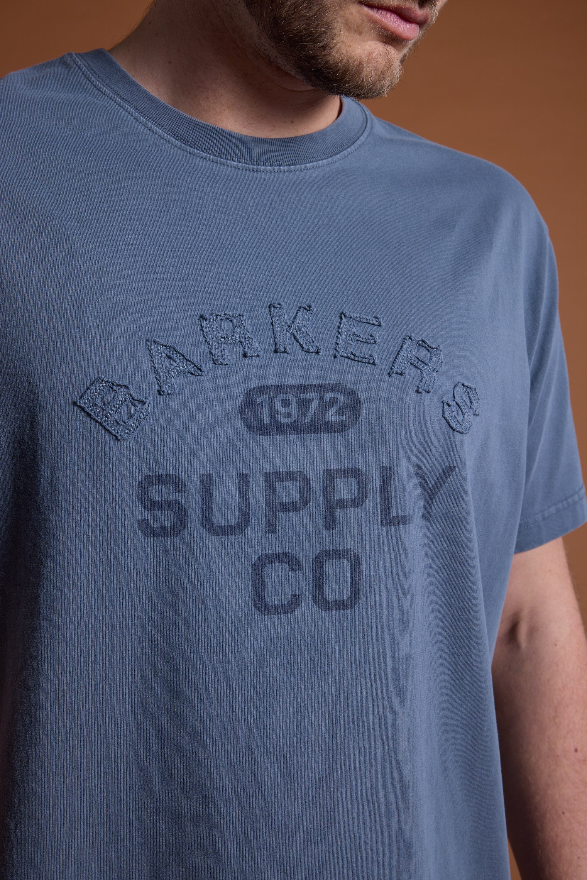 A person wears the Barkers Supply Co GD Print Tee in blue, featuring "Barkers Supply Co 1972" printed and embroidered on the front; their face is partially out of frame.