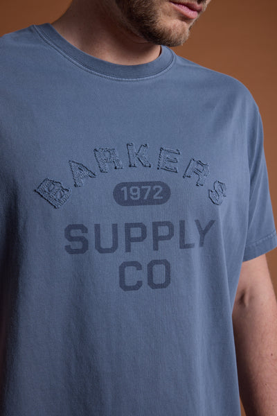 A person wears the Barkers Supply Co GD Print Tee in blue, featuring "Barkers Supply Co 1972" printed and embroidered on the front; their face is partially out of frame.