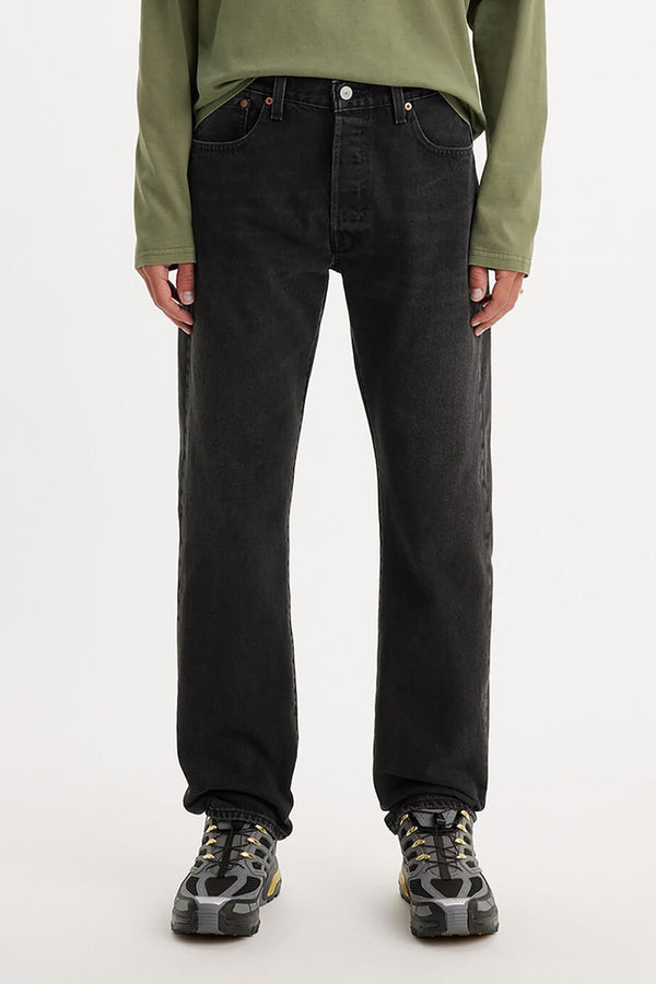 A person stands against a plain white background wearing a light green long-sleeve shirt, gray sneakers, and Levis 501 Original Jeans in black, with their hands relaxed by their sides.