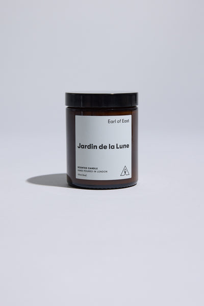 An EoE Jardin de La Lune Candle 170ml by Earl of East rests on a light gray surface, casting a soft left shadow. The brown glass jar’s minimalist white label with black text highlights its hand-poured soy wax quality.