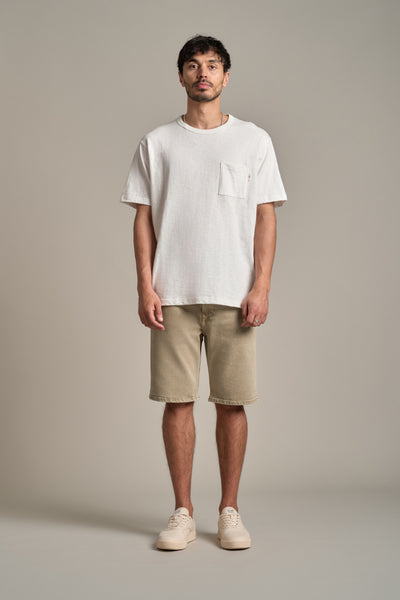 A man faces forward against a plain background, wearing a white short-sleeve t-shirt, Barkers Rover Coloured Denim Shorts in a relaxed fit made from regenerative cotton, and light-colored sneakers.