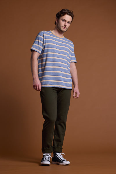 A young man stands against a brown background in the Barkers Chester Stripe Tee—an organic cotton men's t-shirt with blue and beige stripes—paired with dark green pants and black high-top sneakers, looking slightly down in a relaxed pose.