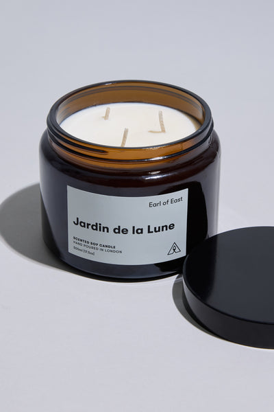 An open 500ml EoE Jardin de La Lune Candle by Earl of East features white soy wax and three wicks in an amber glass jar. The black lid sits beside it on a light gray surface, hinting at notes of tuberose and blackberry leaf.