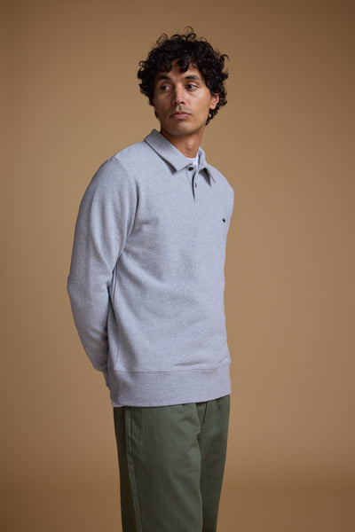A man with curly dark hair wears a Barkers Wallbrook Sweatshirt made from responsibly sourced cotton and green pants, standing against a plain beige background and gazing to the side with his hands behind his back.