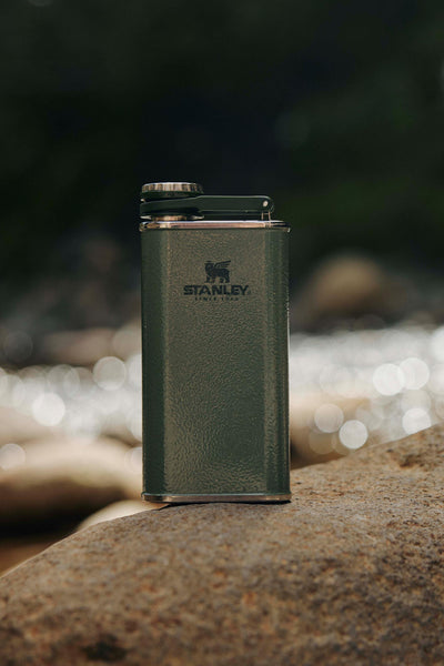 The Stanley Classic Easy Fill Wide Mouth Flask (235ml) by Stanley rests upright on a rock outdoors, making it an ideal adventure companion against a scenic backdrop of water and rocks.