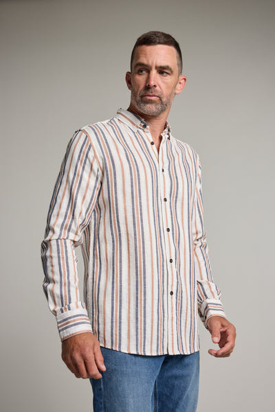 A man with short hair and a trimmed beard models the Barkers Haden Stripe Shirt, featuring vertical beige, blue, and brown summer stripes in a breathable cotton-linen blend, paired with blue jeans as he stands looking slightly to his left.