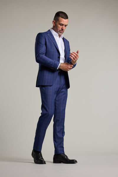 A man in a blue checkered Barkers Paddington Check Suit Trouser and white shirt adjusts his sleeve while standing against a plain light background, wearing black dress shoes and looking down at his arm.