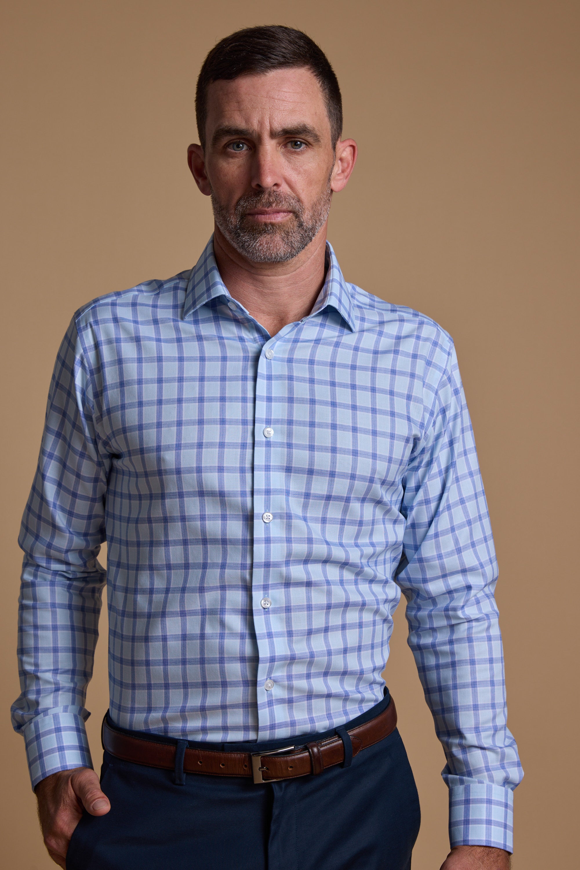 A man with short dark hair and a beard wears a Barkers Bayard Check Business Shirt in light blue and white, paired with dark pants and a brown belt, standing against a plain beige background. The shirt features Eco Easy Care fabric.
