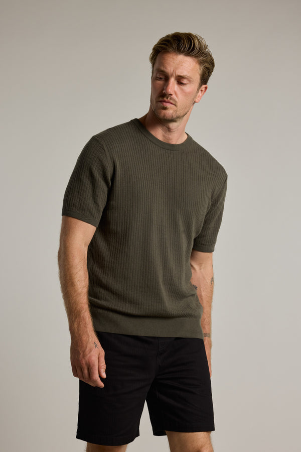 A man with light brown hair and facial hair wears the Barkers Dawn Cotton Knit Tee, a ribbed stripes, short-sleeve, dark green staple shirt, and black shorts. He stands against a plain light background, looking slightly downward with a neutral expression.