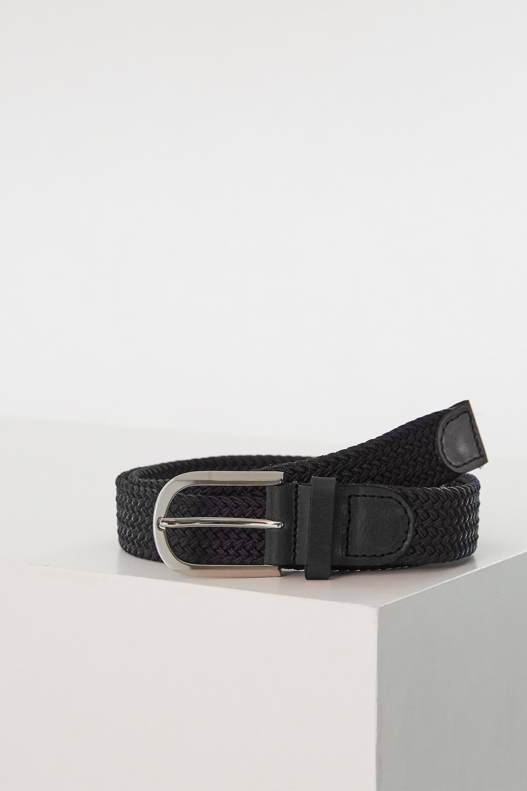 The Barkers Riviera Braided Belt, featuring a black braided design and silver buckle, is coiled on a white surface—ideal for adding a nautical touch to your summer wardrobe in New Zealand.