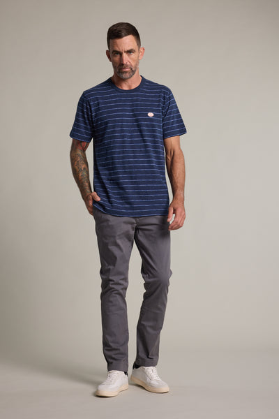 A man with short hair and right arm tattoos stands facing forward, wearing the Barkers Vermont Stripe Tee in navy, paired with gray pants and white sneakers against a plain light background.