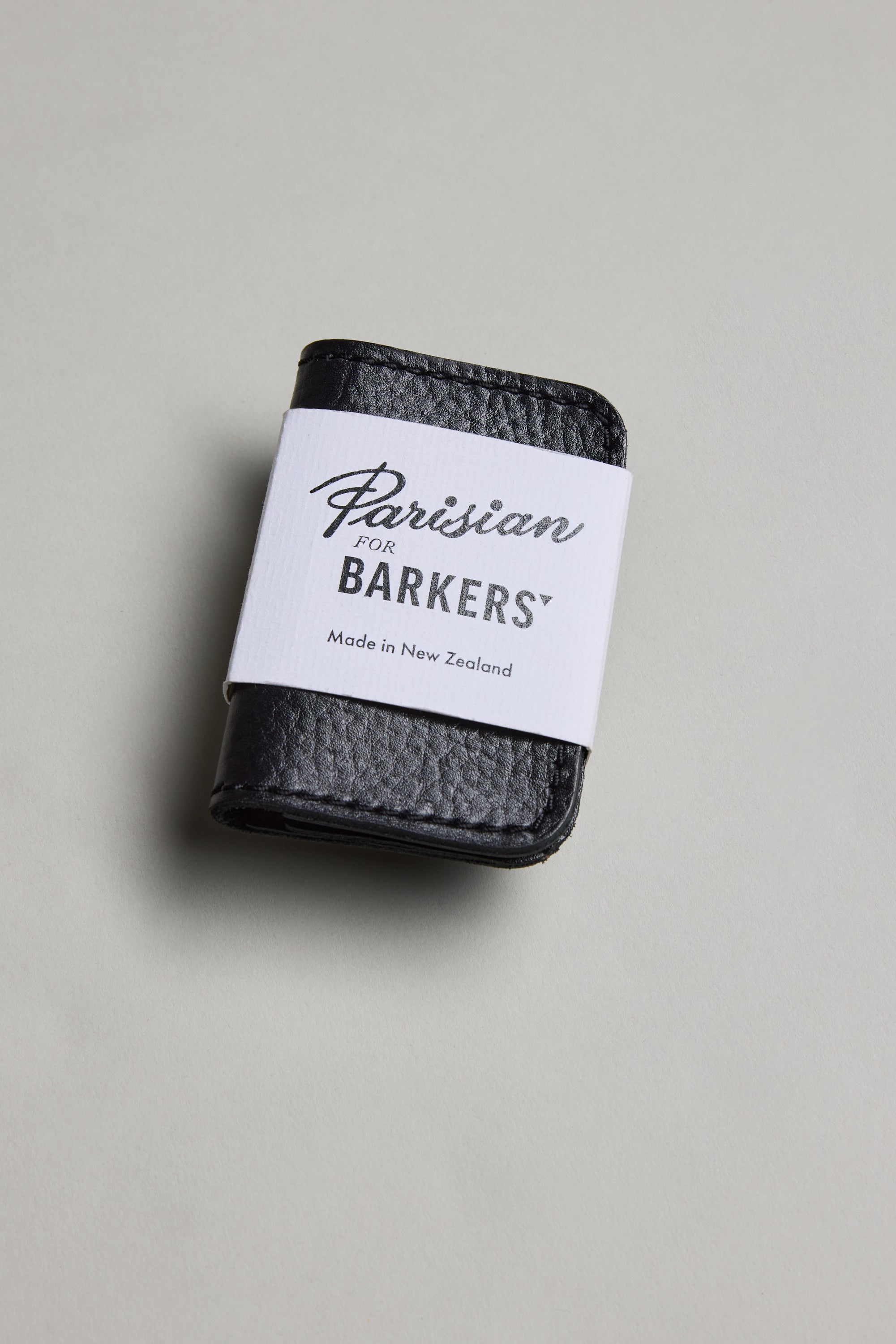 The Parisian Leather Fold Wallet by Parisian, crafted from black New Zealand leather and wrapped with a white paper band, is displayed on a light grey background.