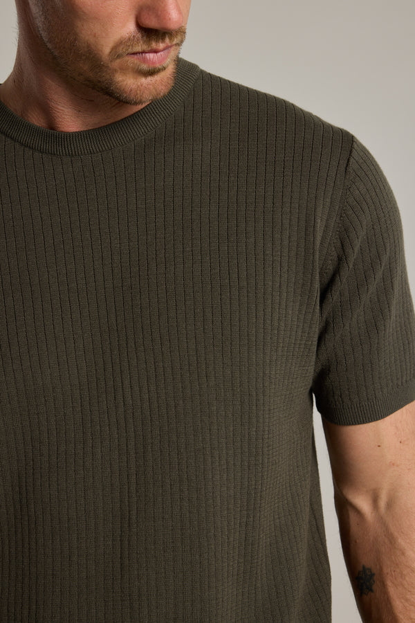 A man wears the Barkers Dawn Cotton Knit Tee, a ribbed, short-sleeve olive-green shirt with a crew neck. Only his lower face, neck, chest, and tattooed right arm are visible against a plain, neutral background.