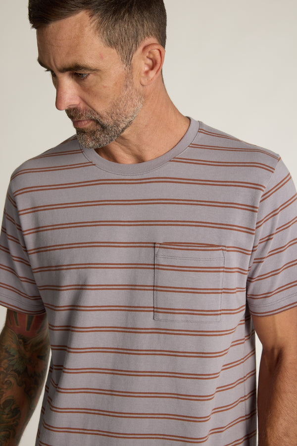 A man with short brown hair and a beard looks down, wearing the Barkers Talbot Stripe Tee—an organic cotton shirt in a boxy fit with horizontal brown stripes and a chest pocket. His left arm has visible tattoos.
