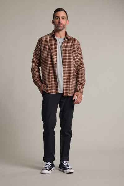 A man with short hair and a beard faces forward in smart casual style, wearing the Barkers Augusta Check Shirt—a tailored fit brown plaid flannel—over a gray t-shirt, with dark pants and black sneakers, one hand in his pocket and a neutral expression.