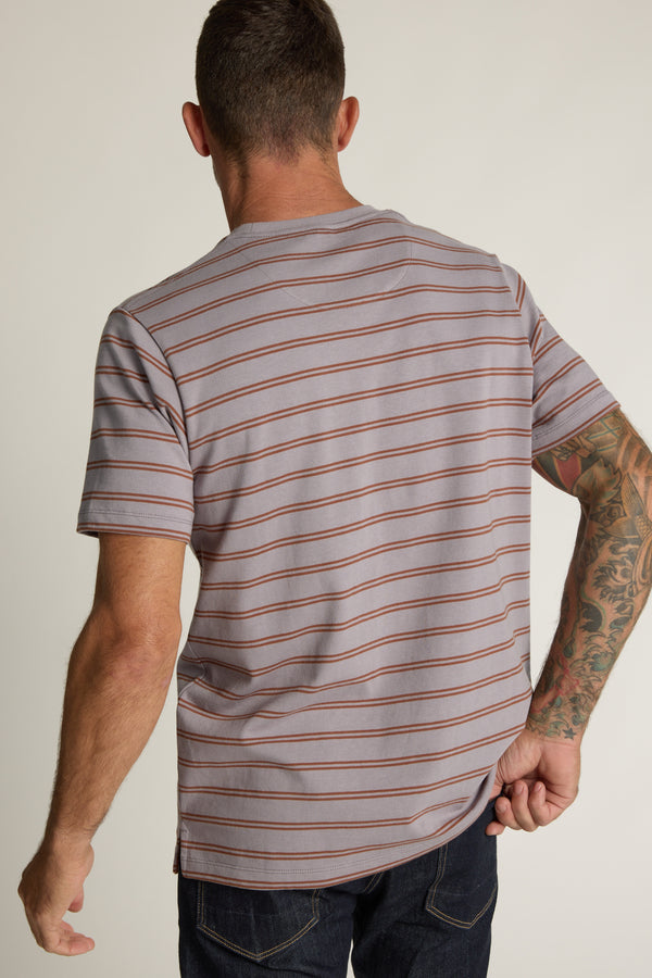 A man with short hair, wearing the Barkers Talbot Stripe Tee—a boxy fit gray top with thin red stripes—faces away. He pulls on the back of his shirt, showing tattooed right arm, and pairs it with dark jeans.