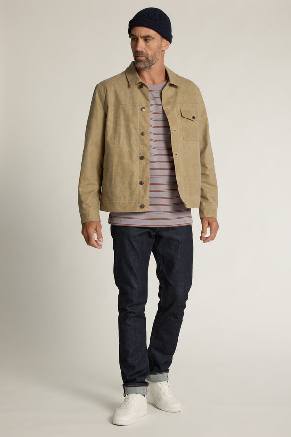 A man wearing a black beanie, tan jacket, Barkers Talbot Stripe Tee, dark jeans, and white sneakers stands against a plain light background, looking to the side.