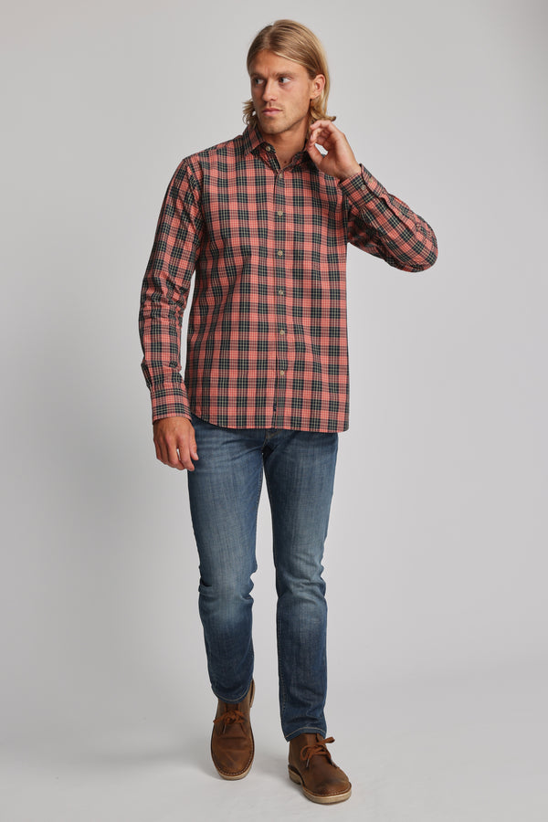 A man with long blonde hair wears the Barkers Formby Long Sleeve Shirt in red and black plaid, blue jeans, and brown shoes. He stands against a plain light gray background, touching his neck and looking to his left.