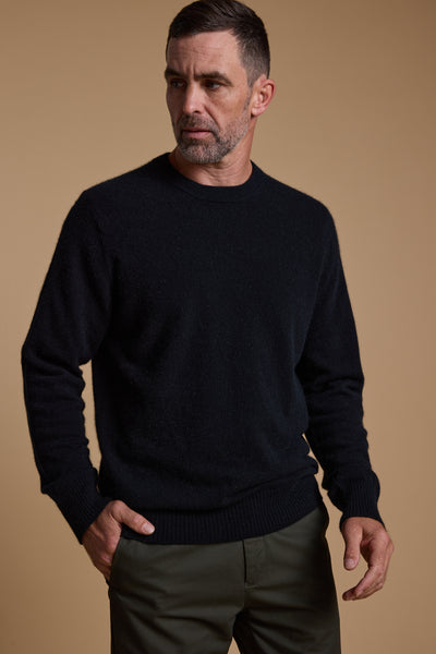 A man with short dark hair and a beard models the Woolyarns Perino NZ Made Perino Crew sweater in black and olive green pants, standing against a beige background with one hand in his pocket, looking slightly to the side.