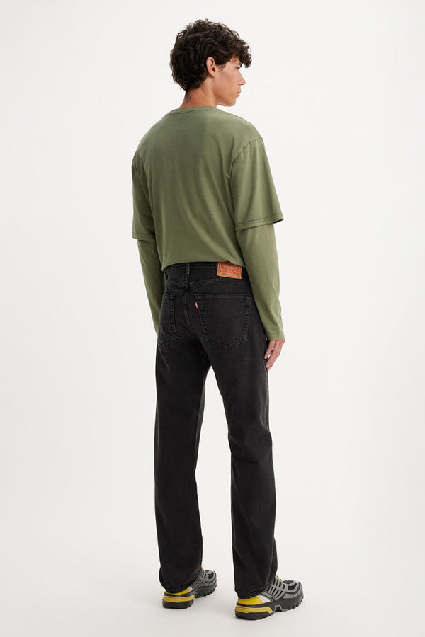 A person stands facing away, wearing a cropped olive green long-sleeve shirt, Levi’s 501 Original Jean by Levis in black, and gray sneakers with yellow accents against a plain light background.