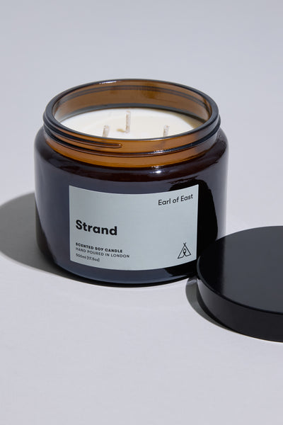 An EoE Strand Candle 500ml by Earl of East in an amber glass jar with a black lid. The label states it's a soy wax candle, hand-poured in London.
