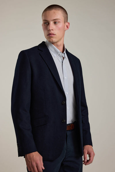 A man stands slightly sideways against a plain background, wearing the Barkers Gilman Woollen Blazer over a light striped shirt and dark pants, looking to his left with a neutral expression.