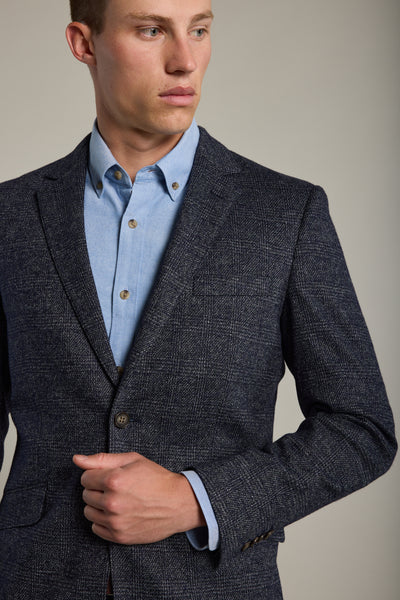A man adjusts his Charlton Knit Blazer by Barkers—a textured blue check design—over a light blue shirt, standing against a plain background and showcasing relaxed tailoring.