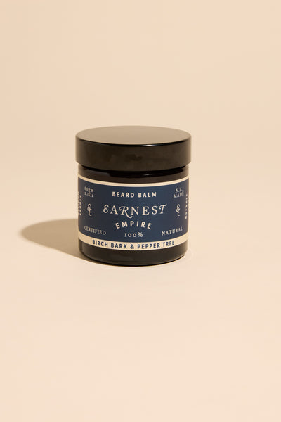 A black-lidded jar of Earnest Empire Beard Balm by Other rests on a beige backdrop. Its label features the Birch Bark & Pepper Tree scent and 100% natural ingredients, detailed in dark blue and white text.