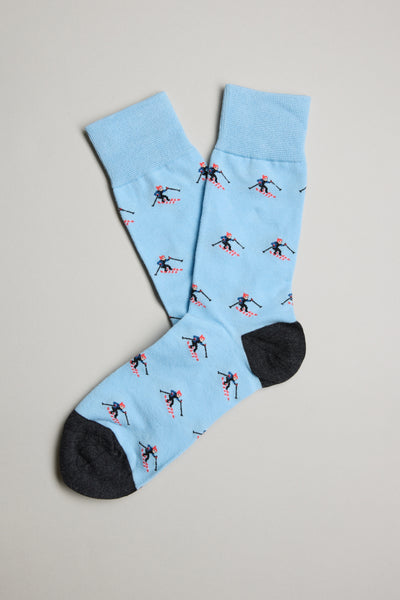The Barkers Glacier Skier Sock in light blue features black toes and heels with a pattern of red and blue skiers. Laid flat on a neutral background, these cotton socks combine comfort and style for ski fans.