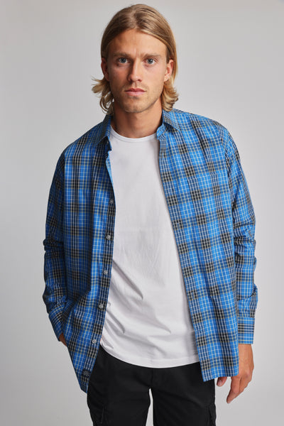 A man with shoulder-length blond hair models the Barkers Edale Long Sleeve Shirt in blue plaid over a white tee and black pants, standing against a plain light gray background.