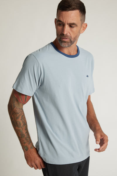 A man with short brown hair and a beard, showing a tattoo sleeve on his left arm, models the Barkers Bailey Peached Tee in light blue with dark blue collar trim. He looks down to his right against a plain background.