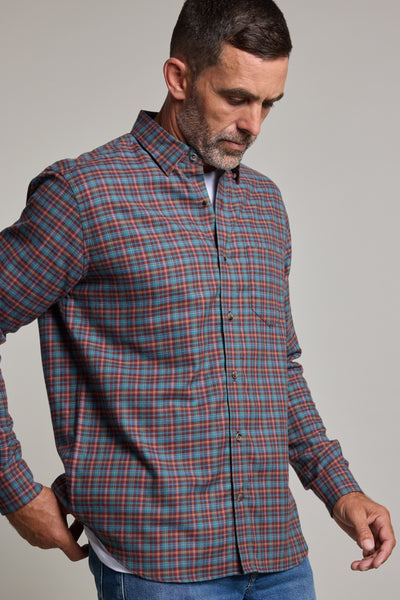 A man wearing the Barkers Dionheck Shirt, a classic fit button-up with long sleeves and a red, blue, and green check pattern, stands against a plain background while looking down and adjusting his shirt.