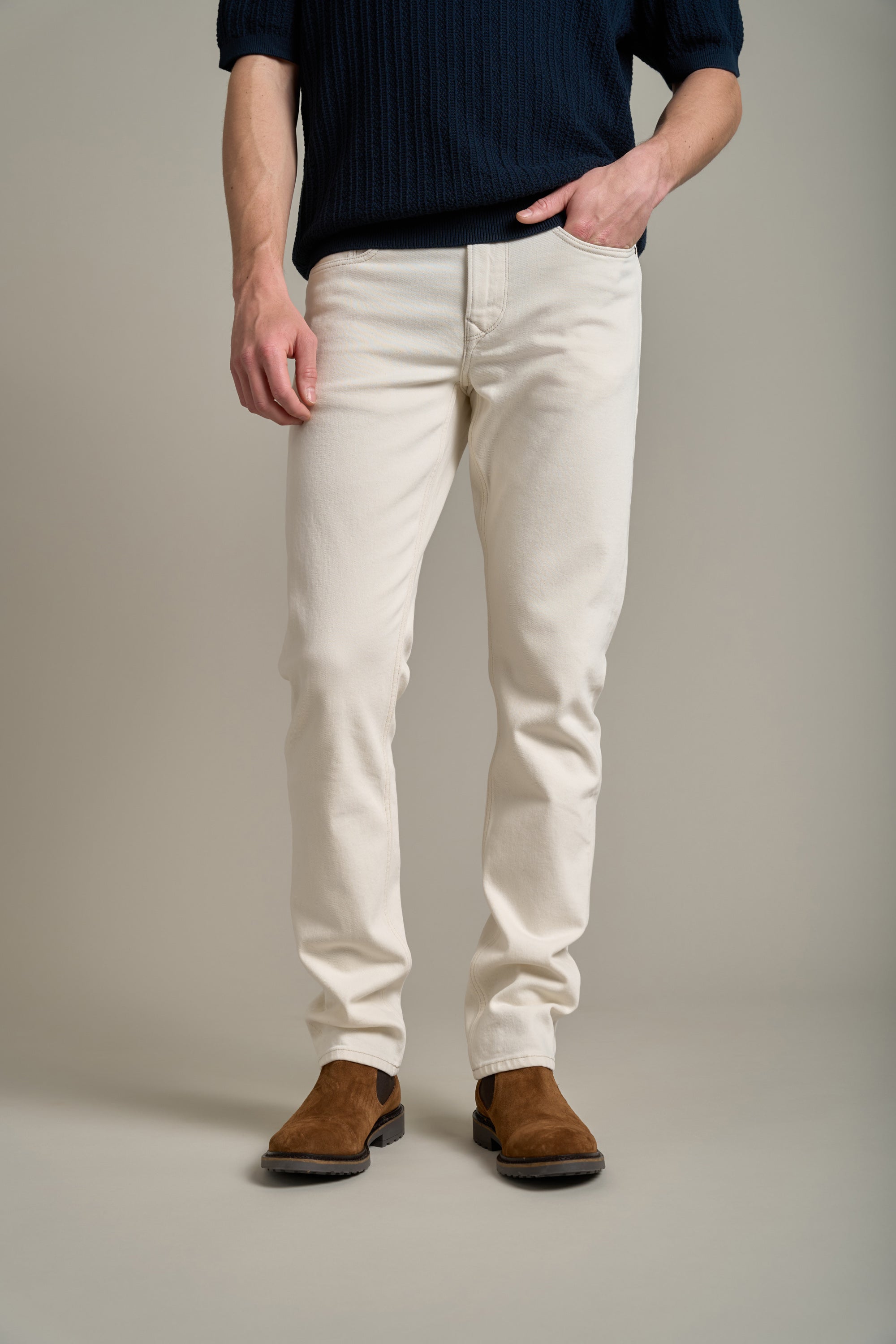 A person stands with one hand in their pocket, wearing Barkers' Nomad Taper Coloured Jean in off-white, a navy knit sweater, and brown suede boots against a plain neutral background. The image is cropped at the shoulders.