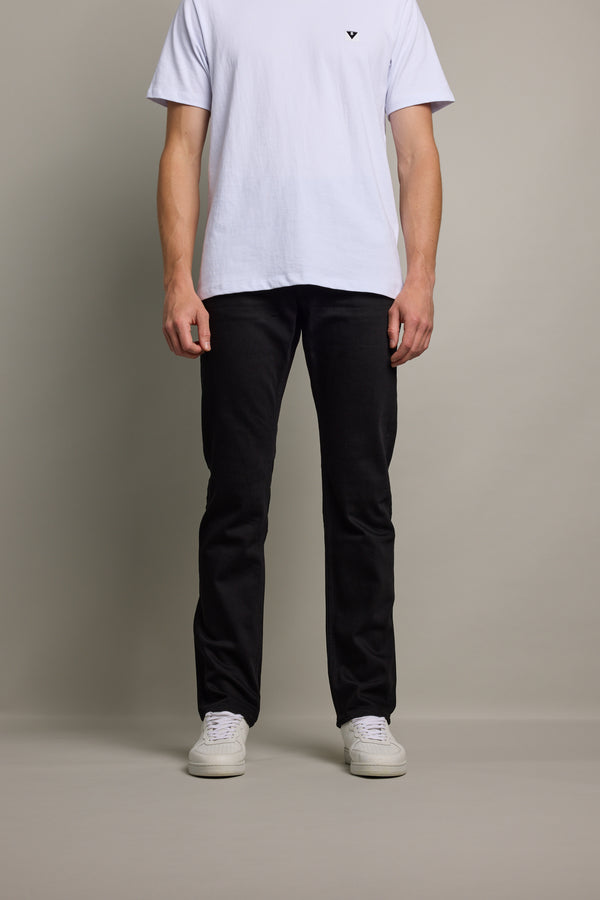A person stands against a neutral background wearing a white short-sleeve t-shirt, Barkers Nomad Denim Taper Jean in black, and white sneakers. The person's head is not visible in the image.