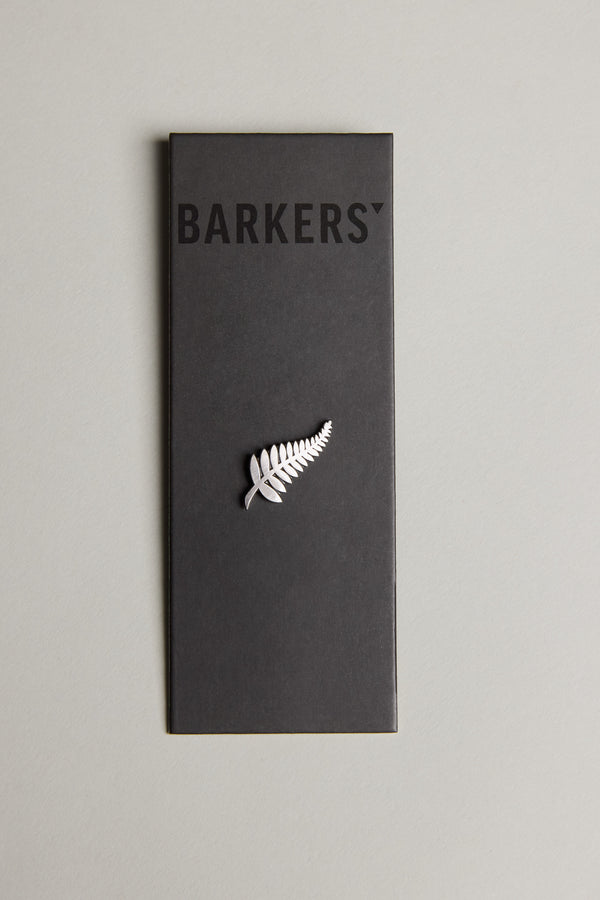 The Classic Fern Lapel Pin from Barkers features a silver fern with a brushed finish, attached to a black card displaying "BARKERS" at the top, all set against a light gray background—a refined accessory.