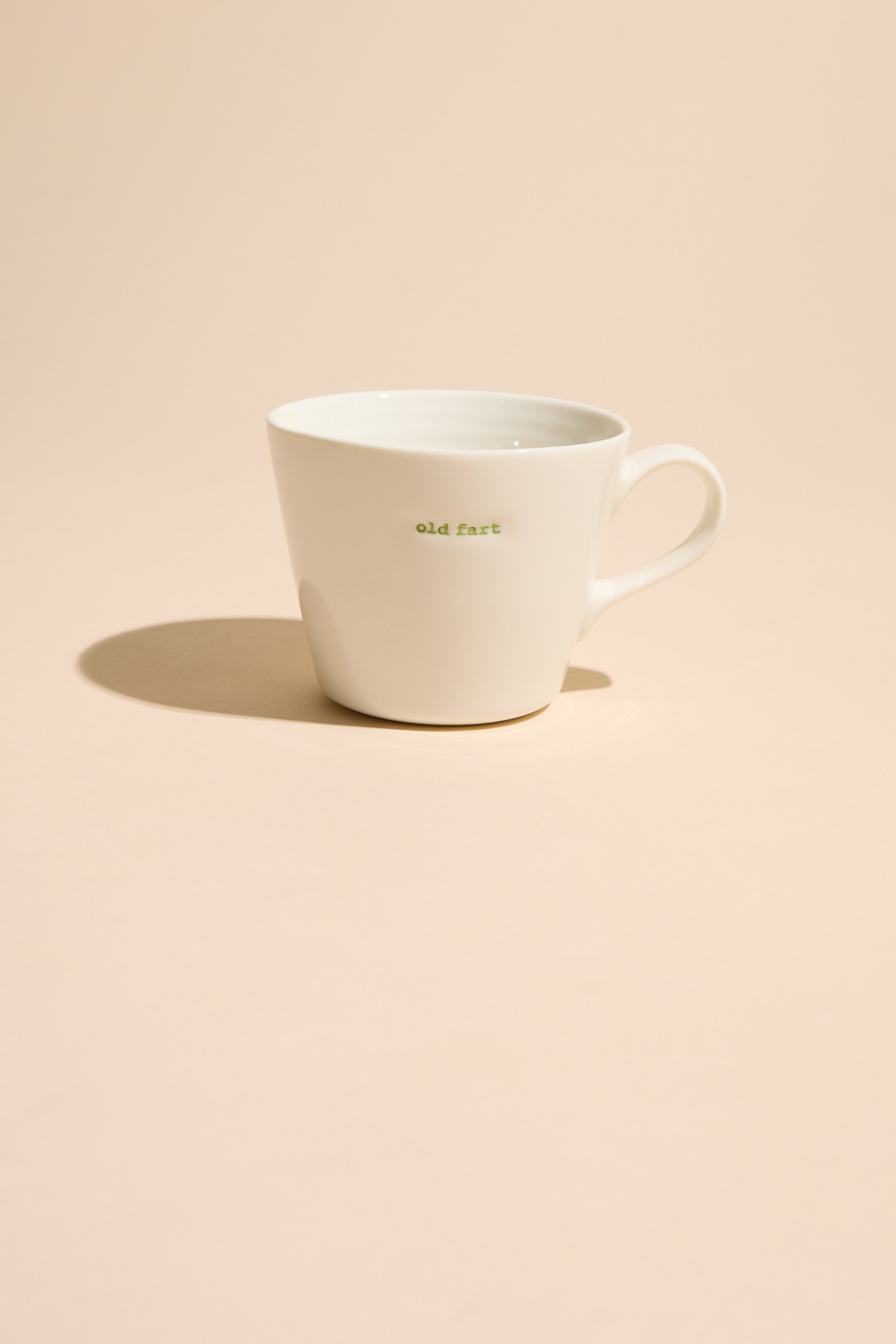 The Bucket Mug - Old Fart by Other is a white ceramic mug featuring "old fart" in small green lettering, set on a beige surface with a soft shadow—an ideal humorous gift for an old fogey.