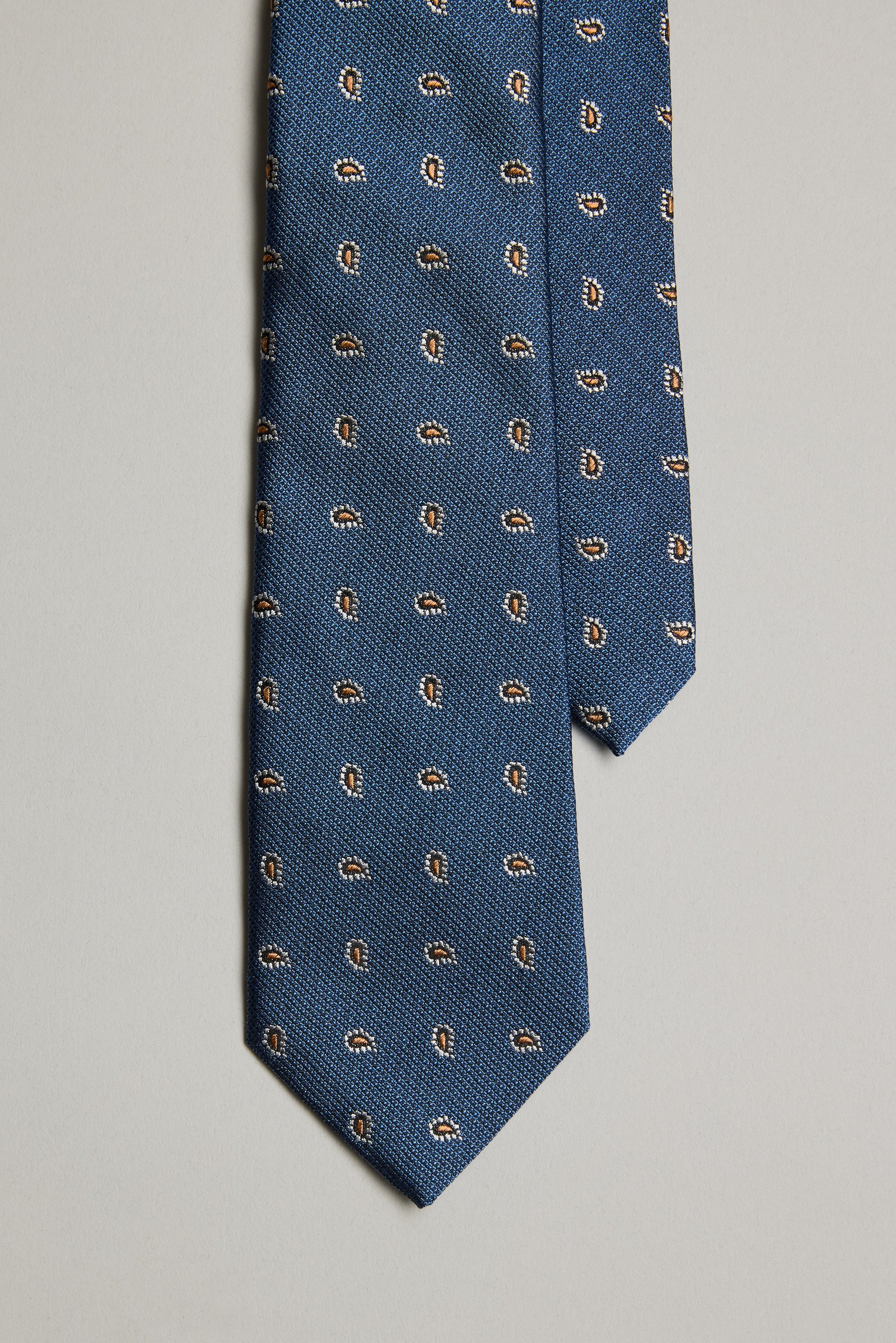 The Kamler Paisley Tie by Barkers is a blue silk tie with a small repeating paisley print in brown, white, and black, shown on a plain light gray background.