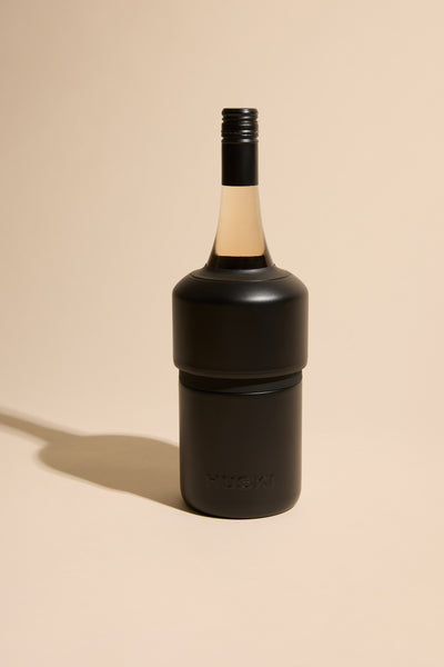 The Huski Wine Cooler by Huski, in black stainless steel, snugly covers the lower half of a wine bottle with a black screw cap, standing upright on a beige background with a soft left shadow to keep your wine cold.
