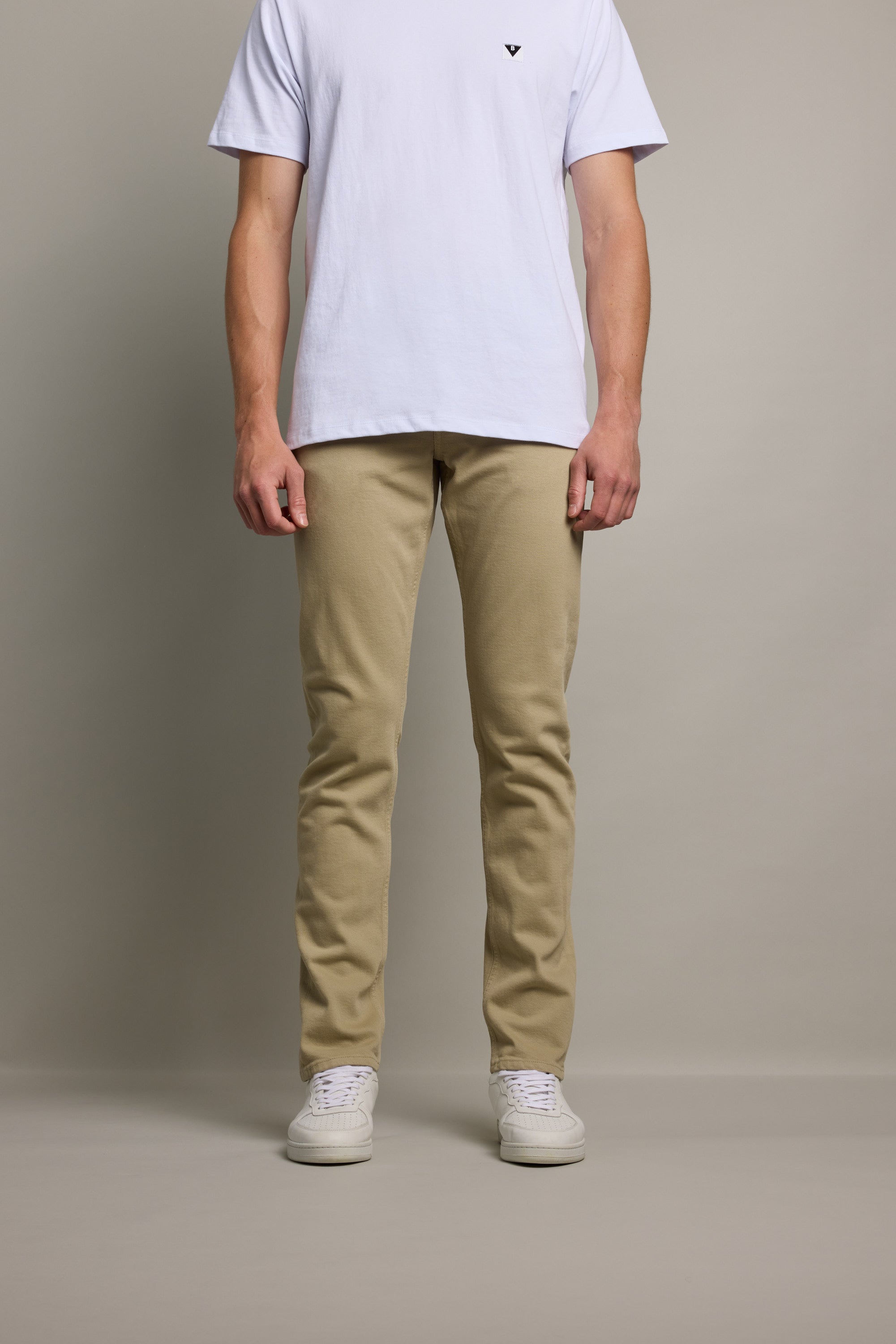 A man stands against a plain gray background, wearing a plain white T-shirt, Barkers Nomad Taper Coloured Jean made from regenerative cotton, and white sneakers. His face is not shown in the image.