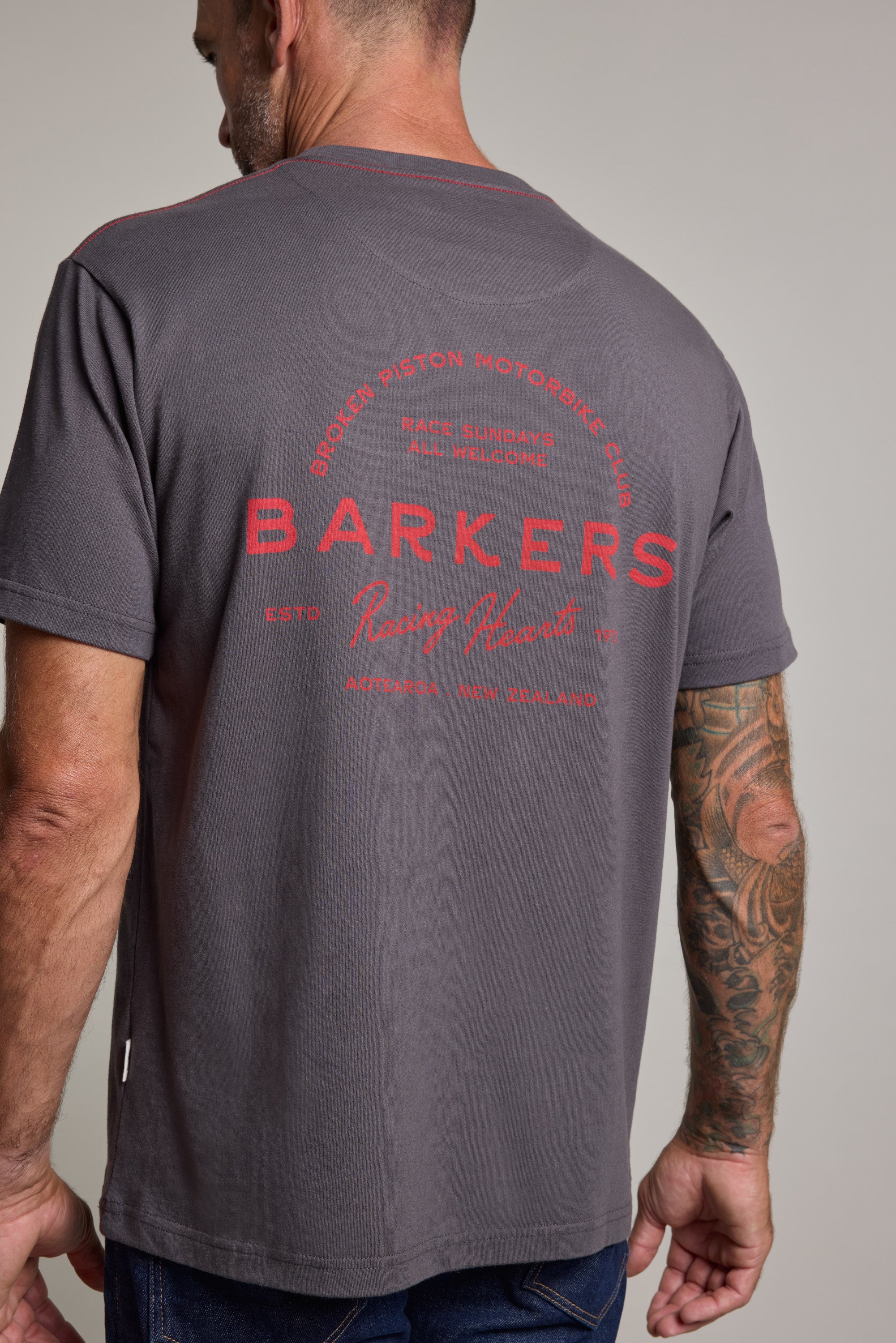 A man with a tattooed arm wears the Barkers Racing Hearts Print Tee, a gray heavyweight graphic shirt with bold red text on the back, standing facing away from the camera.