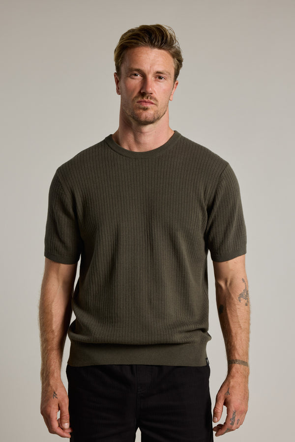 A man with light brown hair, a short beard, and tattoos on his left arm stands against a plain background wearing the Barkers Dawn Cotton Knit Tee in olive green with dark pants. He has a neutral expression.