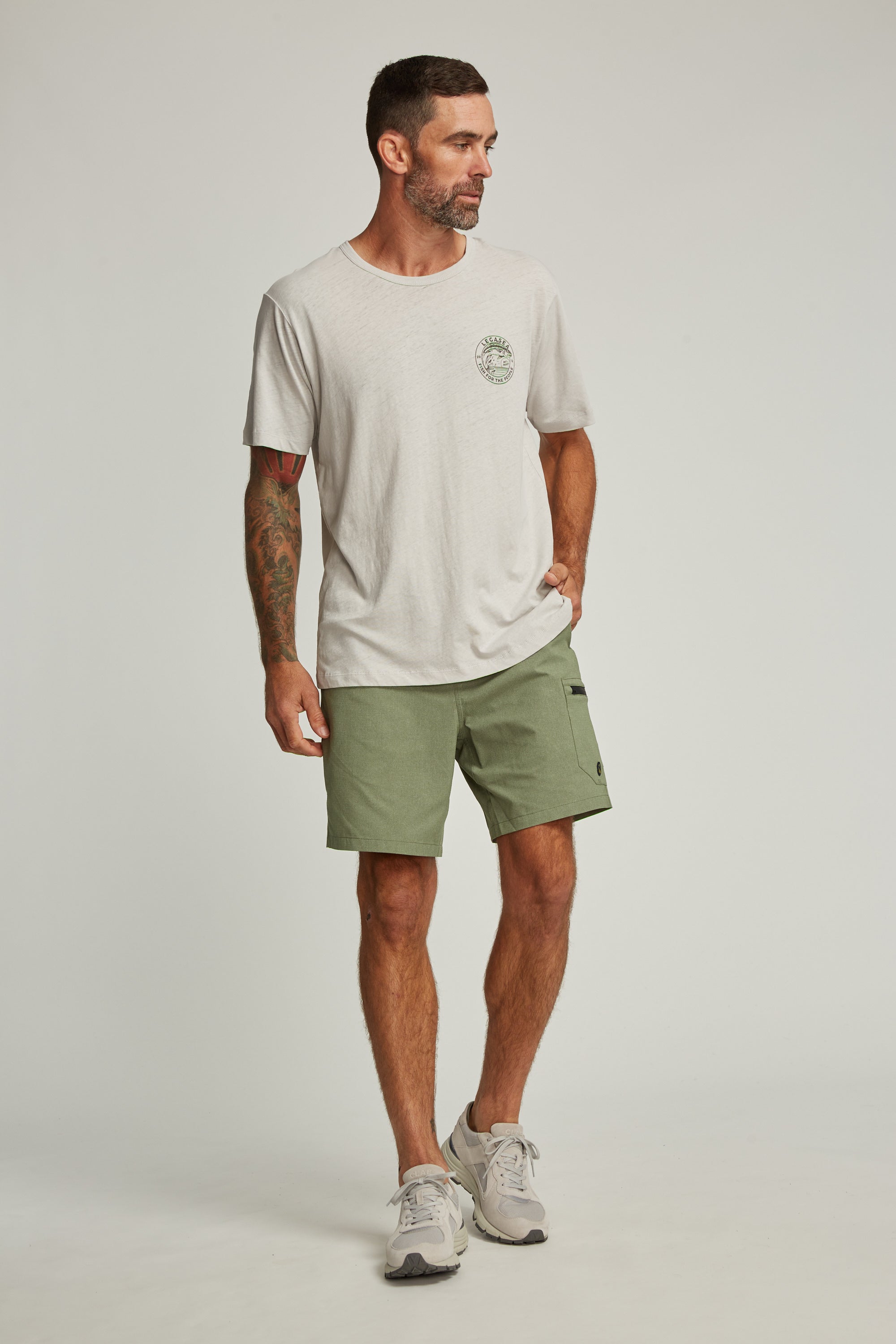 A man with short hair and tattoos on his left arm wears the Legasea Stamp Tee by Legasea, sage green shorts, and gray sneakers. He stands against a plain light background, looking to his left with one hand in his pocket.