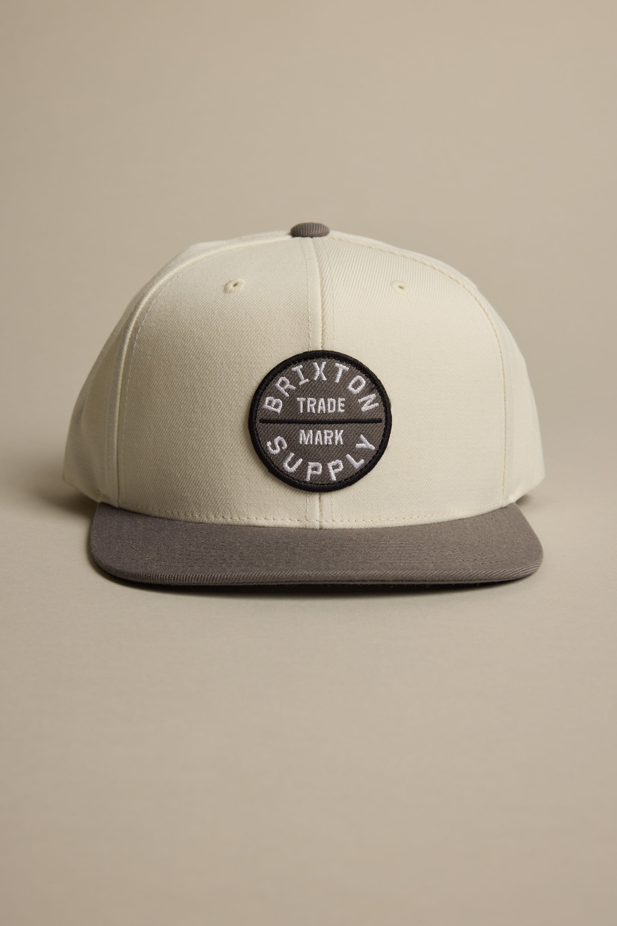 The Brixton Oath III Snapback is a cream hat with a gray brim and a round front patch reading "BRIXTON SUPPLY TRADE MARK" in white. Displayed on a neutral background.