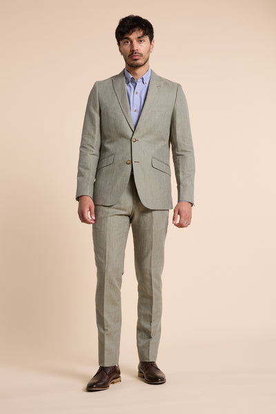 A man stands facing forward in Barkers Pinewood Linen Pant, a light gray breathable linen-blend suit pant, paired with a light blue dress shirt and dark brown shoes against a plain beige background.