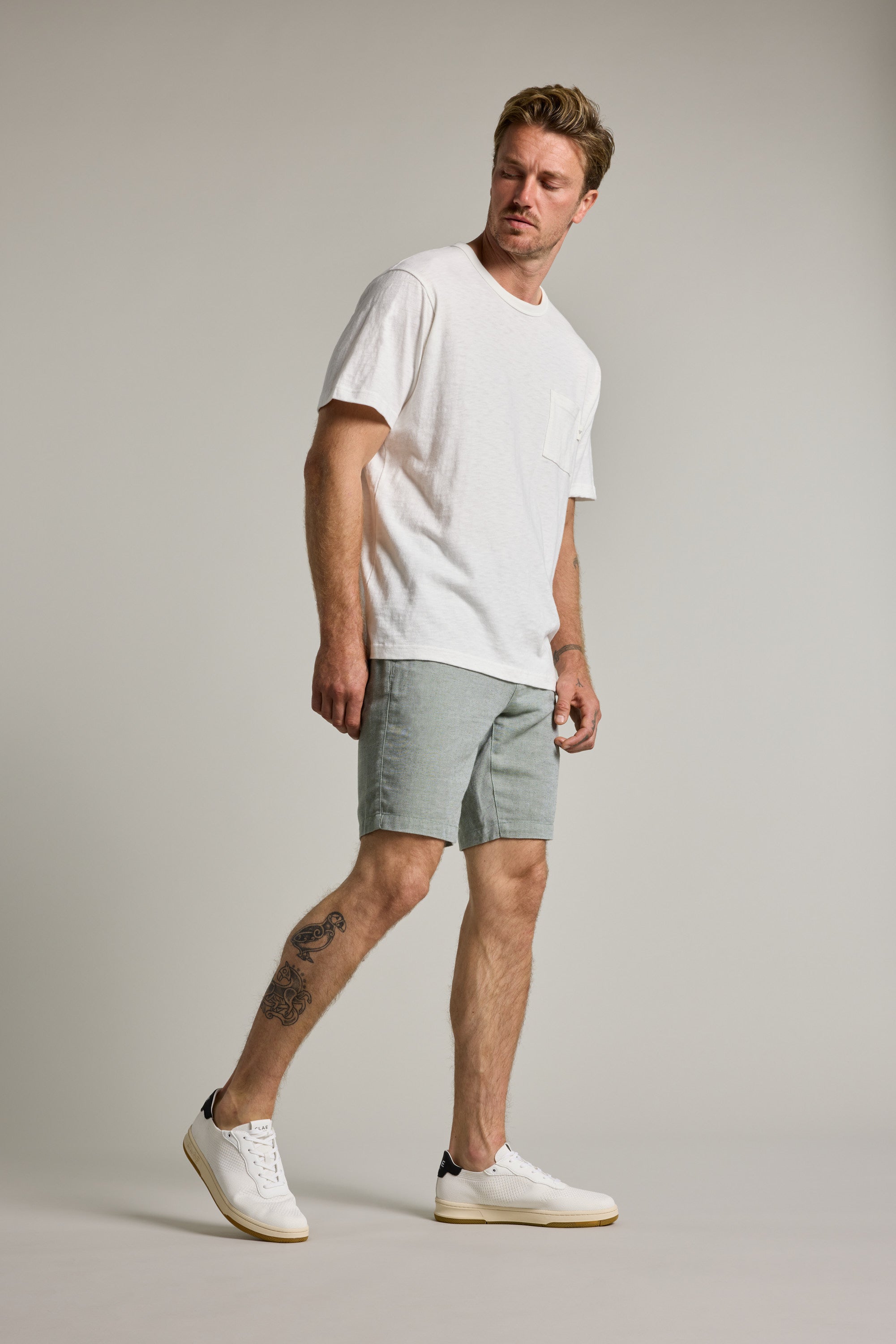 A man with a tattoo on his left leg stands against a plain background wearing Barkers' Elliot Texture Walk Short in light gray, a white T-shirt, and white sneakers. He looks slightly right with a relaxed posture.