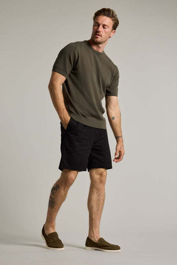 A man with short hair, a mustache, and tattoos on his left leg and right arm wears the Barkers Dawn Cotton Knit Tee—an olive green ribbed crew neck—paired with black shorts and olive slip-on shoes, standing with one hand in his pocket.