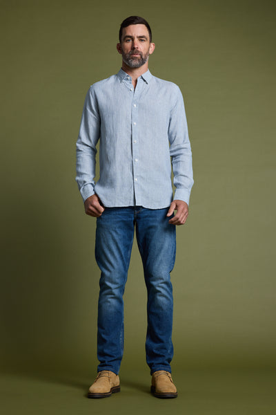 A man with short dark hair and a beard stands against an olive-green background, wearing the Barkers Waterford Shirt—a tailored-fit light blue French linen shirt—paired with blue jeans and tan suede shoes, ideal for summer.