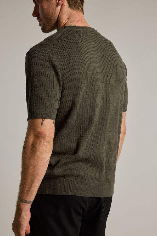 A man is seen from behind wearing the Barkers Dawn Cotton Knit Tee, an olive green, short-sleeve sweater with ribbed stripes and a crew neckline, paired with black pants. Tattoos are visible on his right arm as he stands against a light background.